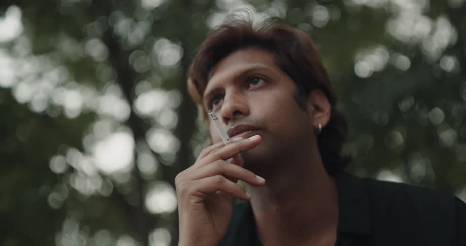 Man with brown hair earring and black shirt smokes a cigarette under the trees