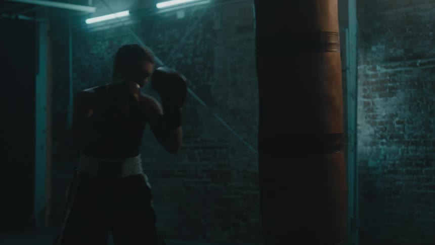 A female boxer trains in a dimly lit room by striking a punching bag