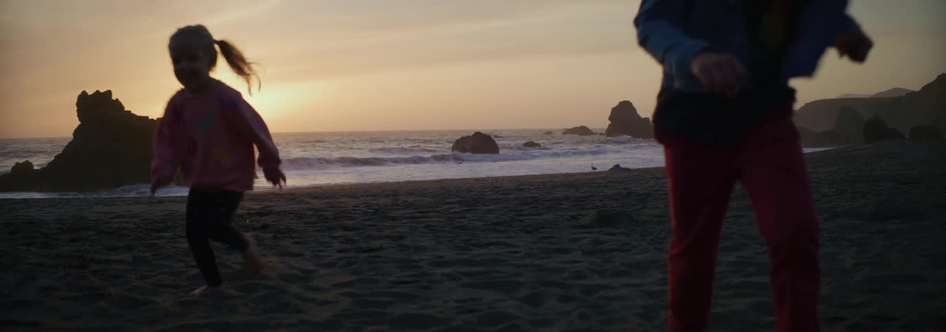 Two children are running on a beach as the sun sets over a rock formation in the water