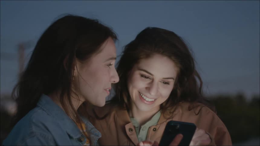 Two women with denim jacket brown jacket and wavy hair look at smartphone laughing outdoors at dusk on a rooftop