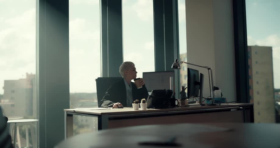 Businesswoman with short fair hair black blazer jacket and blue shirt sits in chair drinking coffee from cup in the office