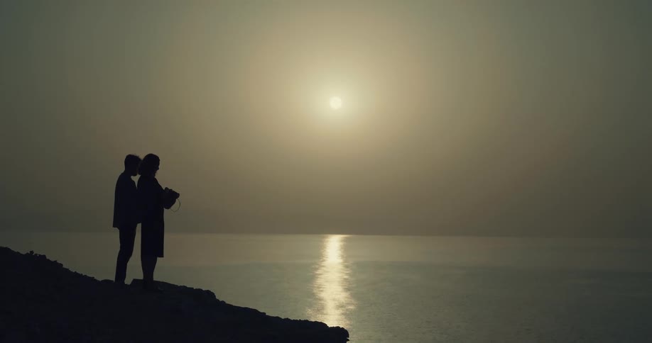 The silhouette of two people standing on a rock throwing ashes into the sea