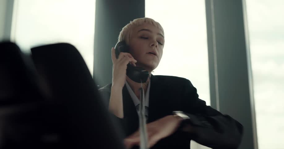 Businesswoman with short hair black blazer jacket and shirt sits at desk using smartphone and answering phone in the office