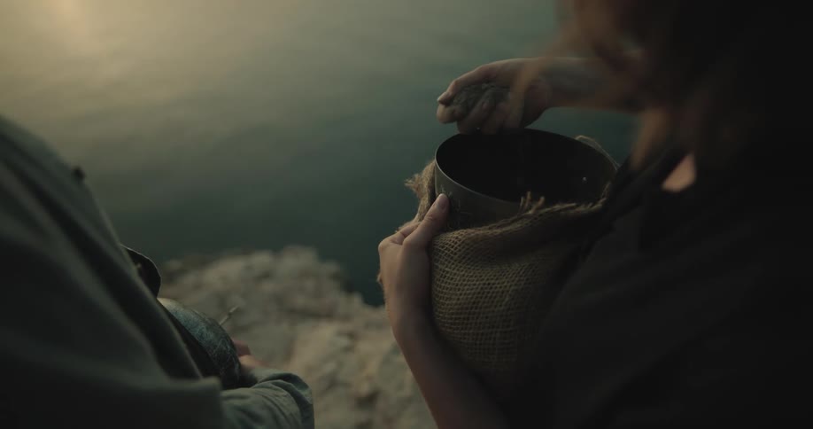 A woman throws handfuls of ashes from a bowl as she stands with a man on the coast of a body of water