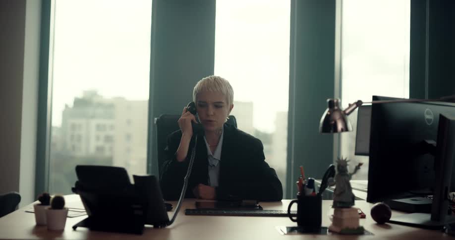 Businesswoman with short hair blazer jacket and blue shirt sits at desk answering phone in the office