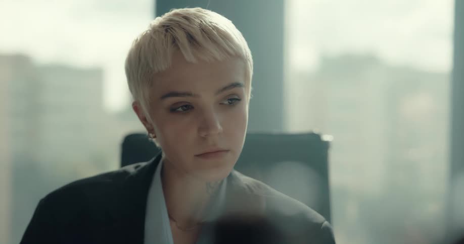 Businesswoman with short fair hair black blazer jacket and blue shirt sits at desk using computer in the office