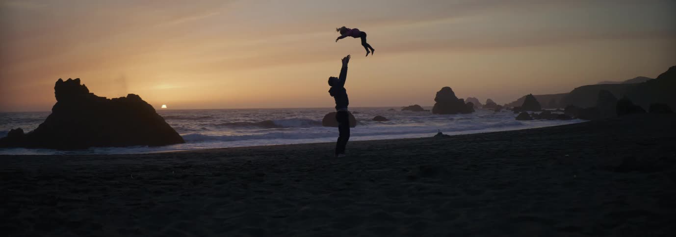 Silhouettes of a man playing with his little girl and tossing her into the air on a beach as the sun has set
