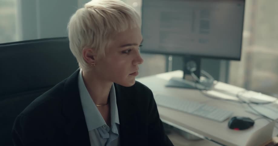 Businesswoman with short fair hair black blazer jacket and blue shirt sits in chair drinking coffee from cup and anwering phone in the office