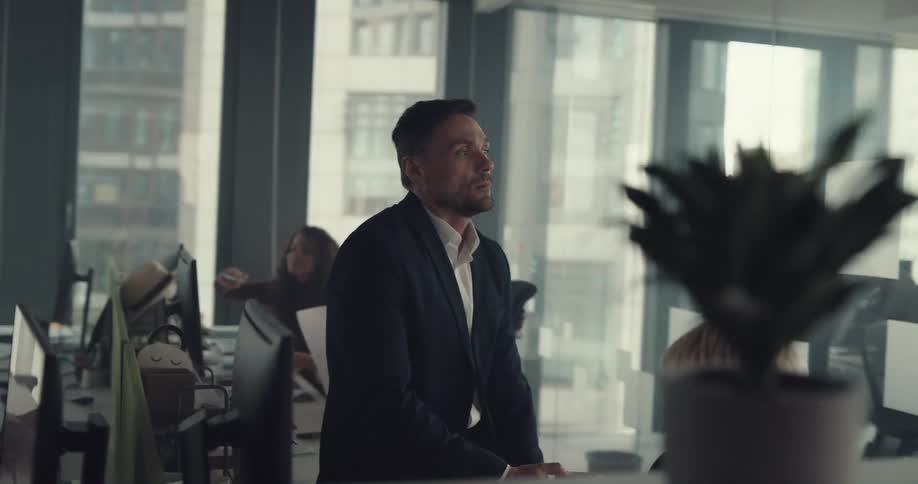 Boss sighs and checks his watch while sitting on desk near employee talking on the phone