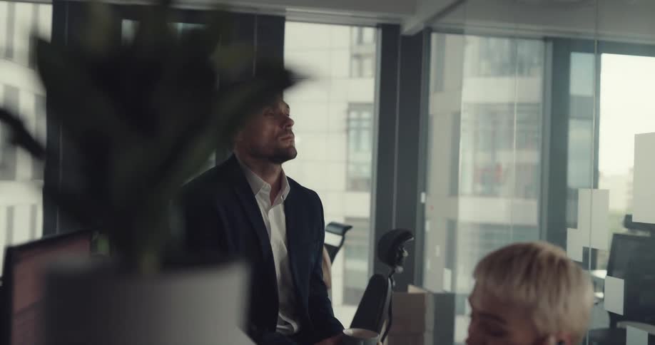 A man sits silently drinking from a mug while a woman quickly works beside him in an office