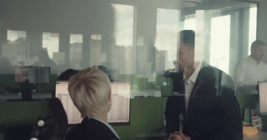 Boss sits at employees desk talking to him and others in office