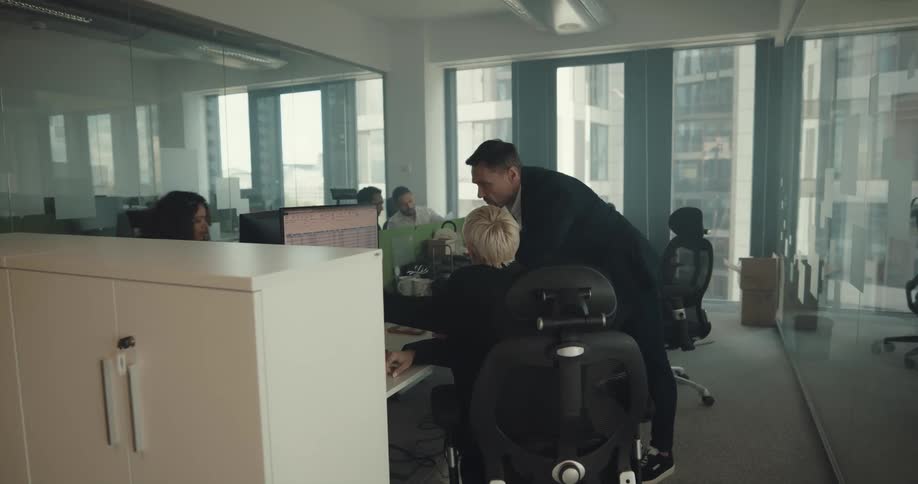 A man walks through a busy office and stops at a desk to talk to a woman