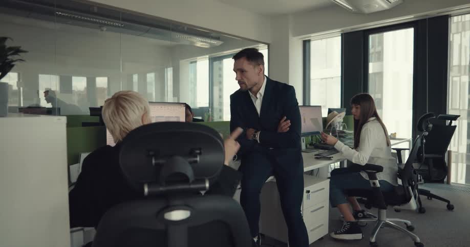 A man in an office walks to a womans desk waves at other employees and sits on the desk and listens to the woman as they talk