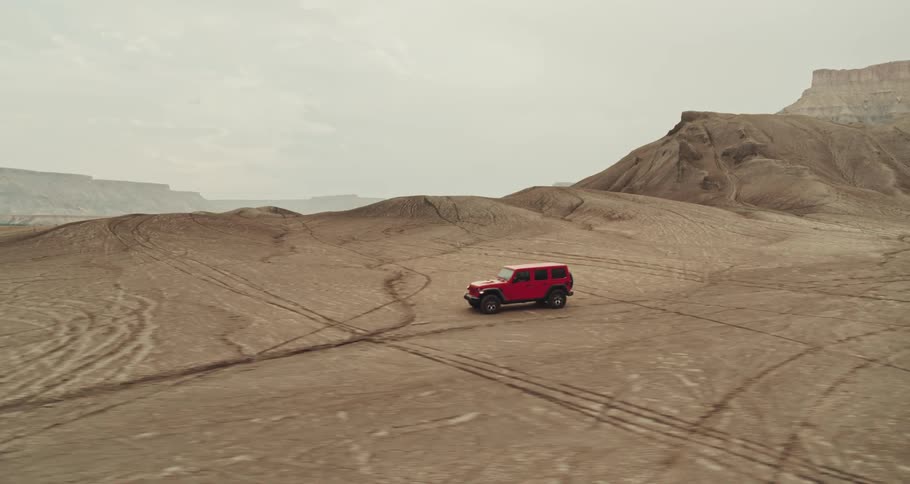 A red truck travels across the desert with mountains and mesa in the distance under a hazy sky