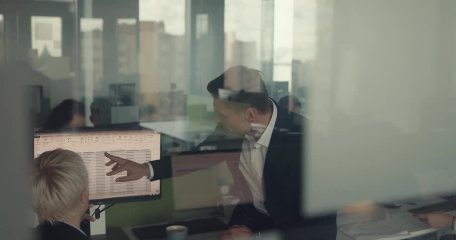 Boss sits on desk discussing data on monitor with employee