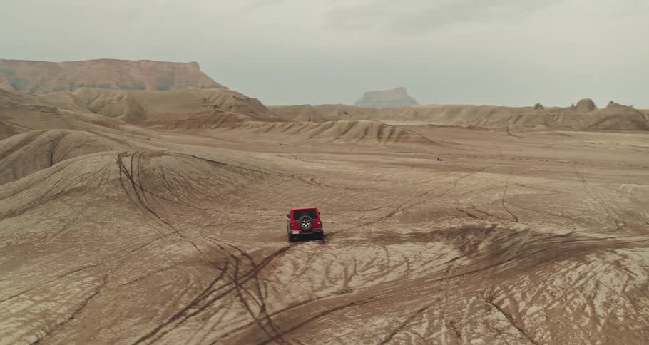 A red truck travels across the desert with mountains and mesa in the distance under a hazy sky