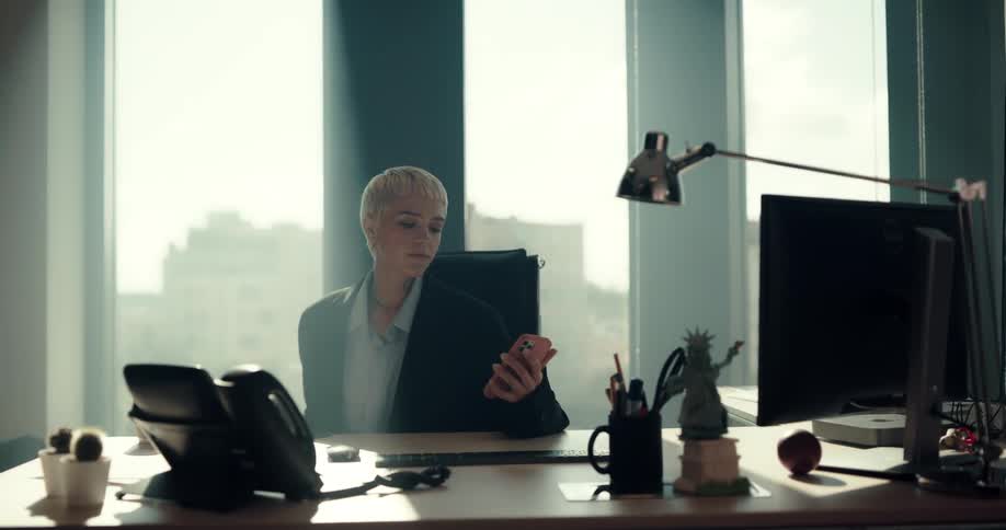 Woman sitting at a desk in her office puts down her phone and starts working on a computer