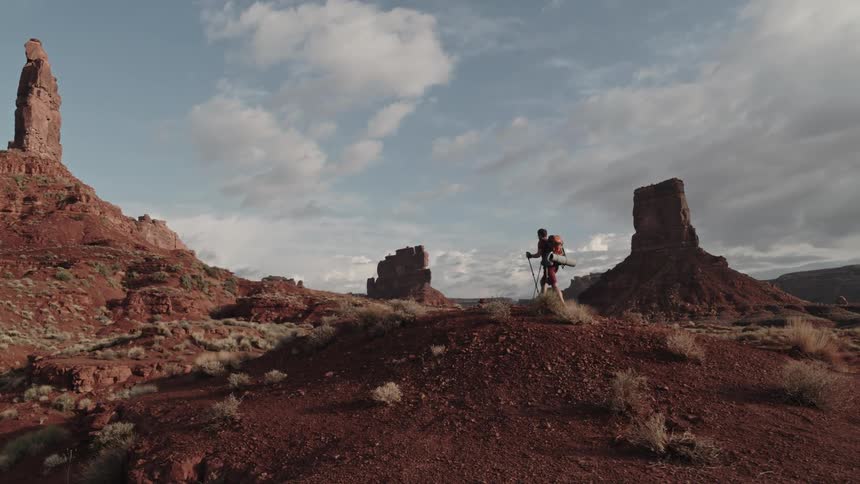 A man uses trekking poles to hike to the top of a hill in the badlands during the day