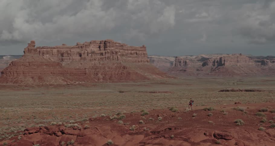 A man is standing on a hill with his foot on a large rock and buttes and mesa are in the distance under a cloudy sky