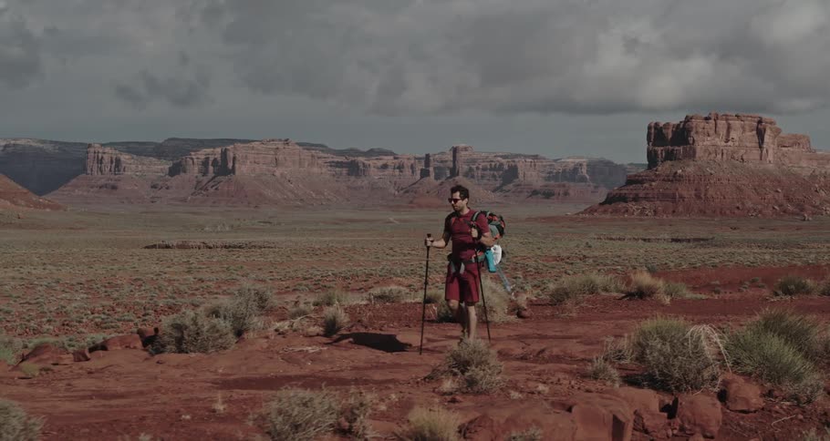 A man is hiking through the desert badlands with trekking poles and there are buttes and mesa in the distance under a cloudy sky