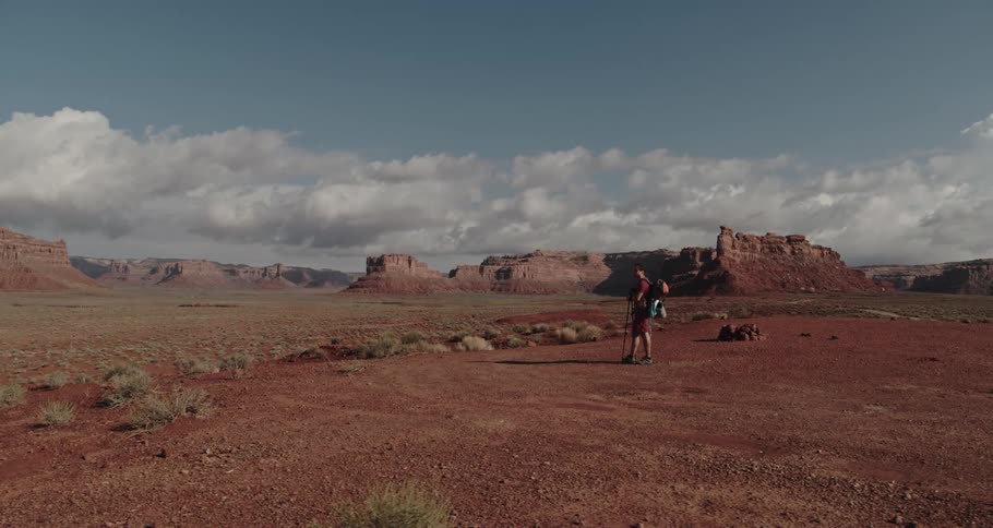 A hiker is standing in the desert badlands holding trekking poles and there are buttes in the distance under a cloudy sky