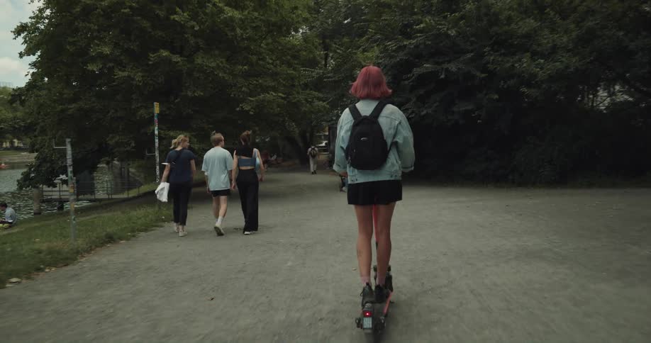 A young woman rides an electric scooter down a park pathway around other people walking