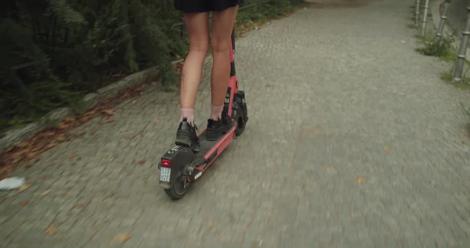 A woman wearing a backpack is riding a pink electric scooter on a park sidewalk