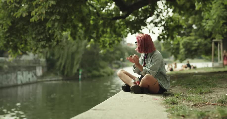 A teenage girl with pink hair and sunglasses is sitting in a park near a stream and she rolls a joint