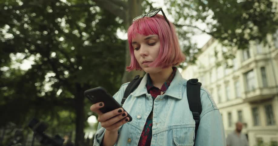 A teenage girl with pink hair and sunglasses on her head is outside near trees using her cell phone