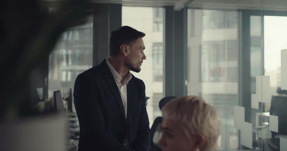 Boss frowns and drinks coffee while sitting next to employee studying computer monitor