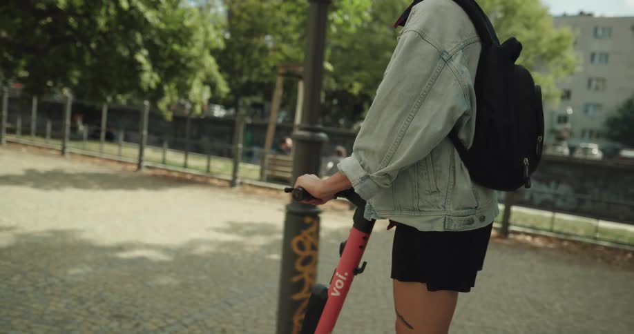 A teenage girl with pink hair and a backpack is looking around as she rides a scooter through the park