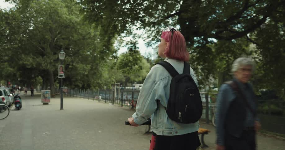 A young woman looks around at her surroundings as she rides a scooter down a walkway on a waterfront during the day