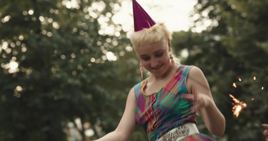 A pair of women wearing party hats wave sparklers while dancing outdoors during the day