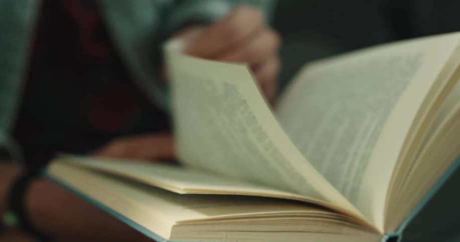 A girl wearing black nail polish is sitting down holding a book and turning pages as she reads