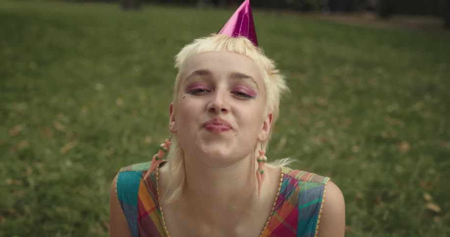 A woman smiles and makes faces as she sits in the grass wearing a party hat