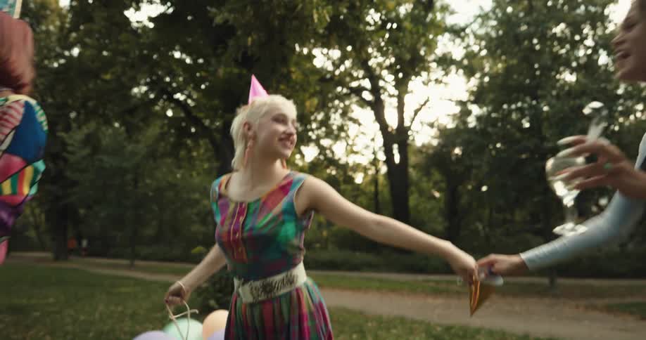 Three teenagers dance around in a park with balloons party hats and wine while one blows bubbles