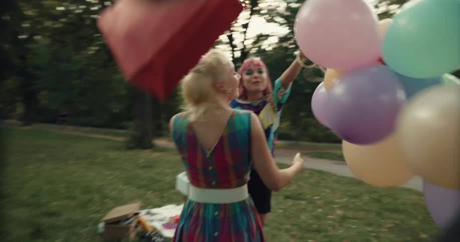 A group of three young women all hug excitedly outside with party bags balloons and gifts