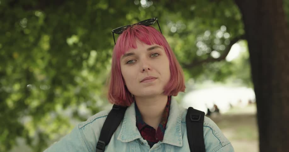 A teenage girl with pink hair and sunglasses on her head is outside near trees and smiling