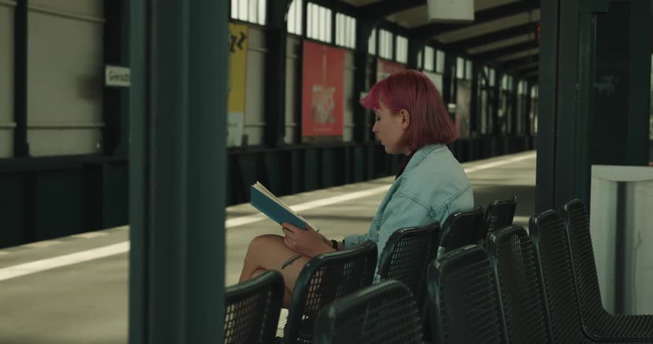 A teenage girl is sitting in a chair at a train station reading a book
