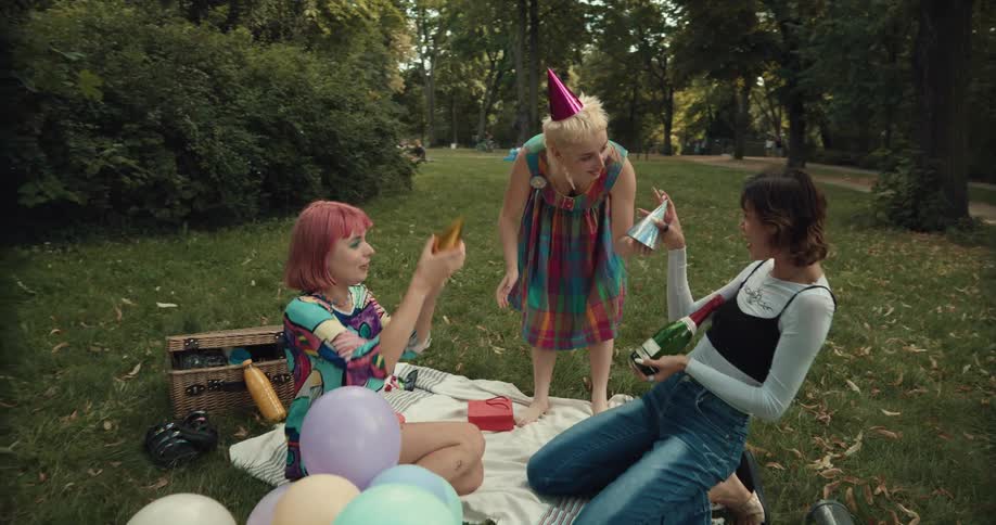 A group of women put on birthday hats as they sit on a blanket in a park with a bottle of wine