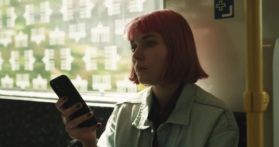 Young woman with pink hair sitting on seat inside a moving train looking at her phone