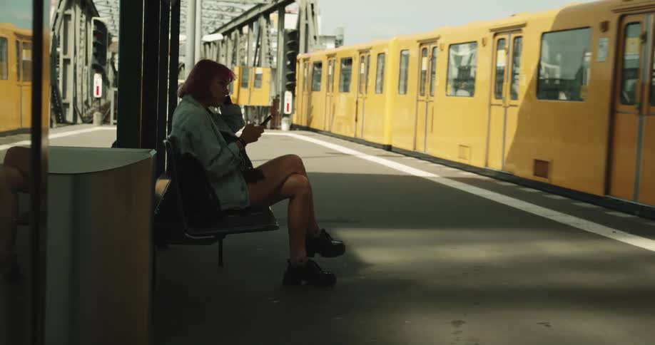 A teenage girl is sitting in a chair at a train station using her phone as trains travel past her