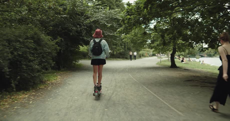 Woman with pink hair wearing shorts rides an electric scooter in the park at daytime