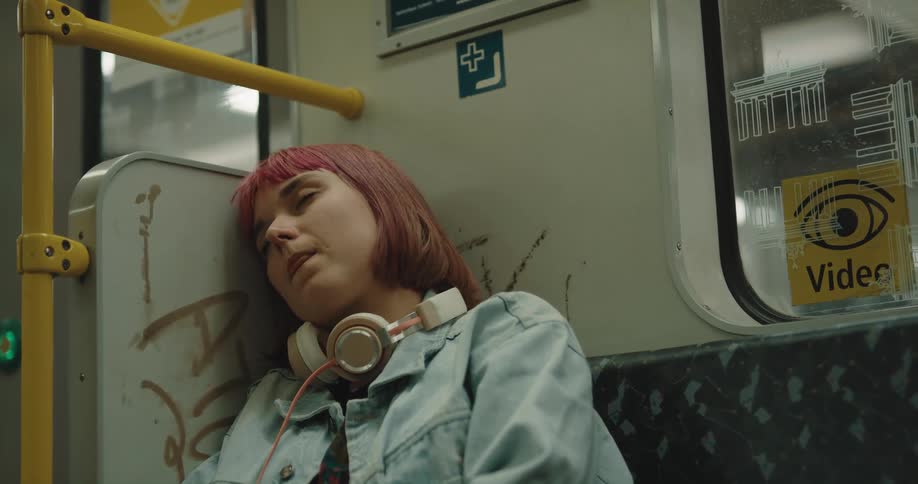 A teenage girl with pink hair and headphones around her neck is sleeping on a train