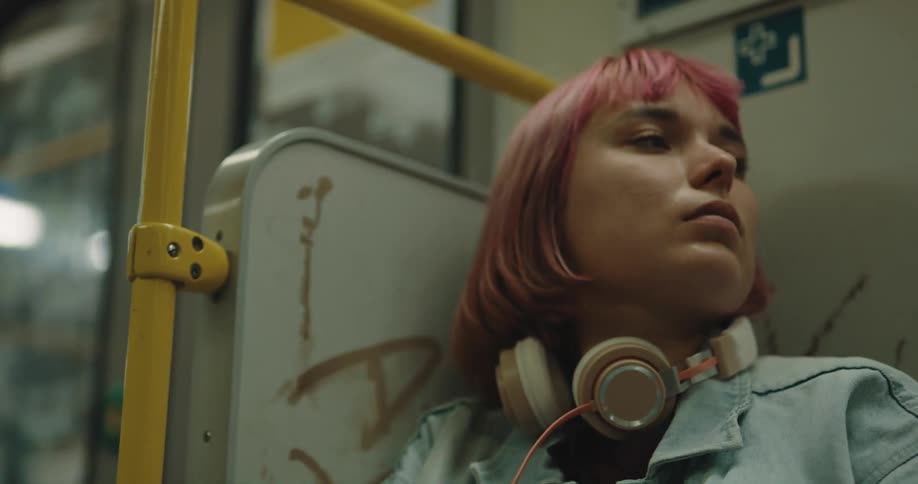 A teenage girl with pink hair and headphones around her neck is sleeping on a train
