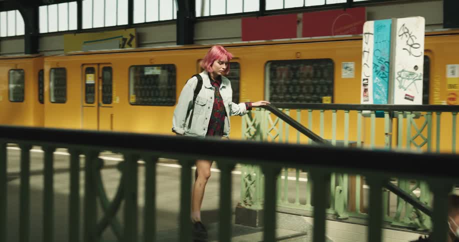 A teenage girl with pink hair and a backpack gets off a train and hurries down the stairs