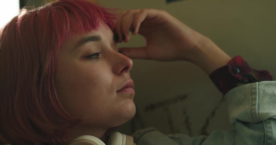 A teenage girl with pink hair and headphones around her neck is sleeping on a train