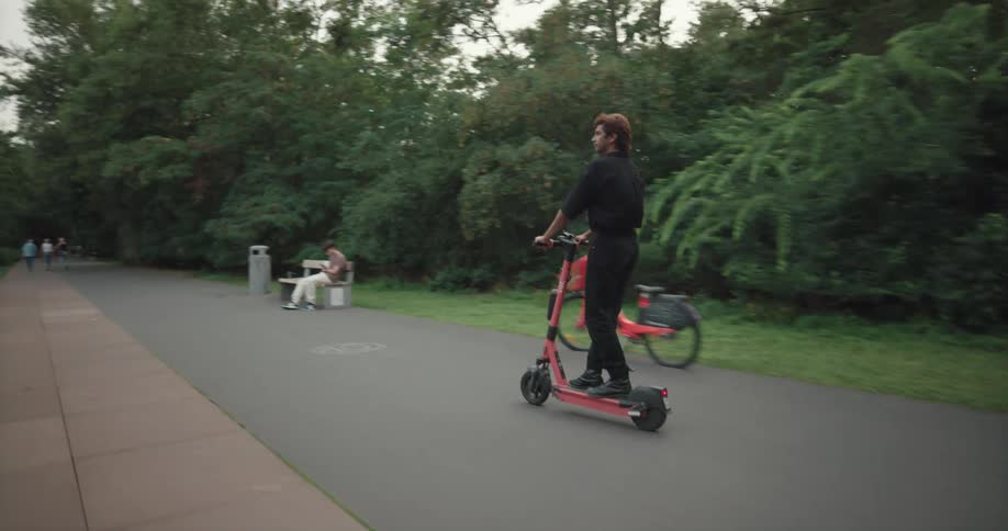 A man rides a scooter down a path in a park past a teenager sitting on a bench by a bike
