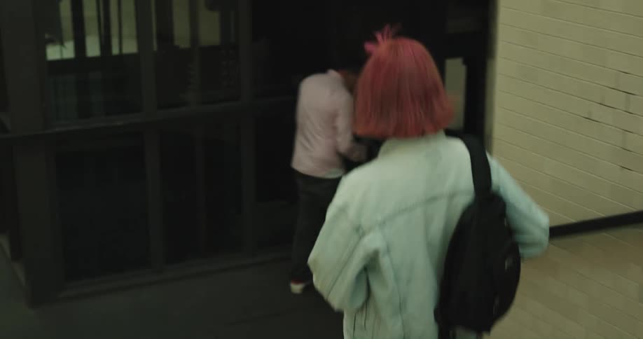 A teenage girl with pink hair and a backpack gets off a train and pulls her phone out of her pocket as she walks down the stairs