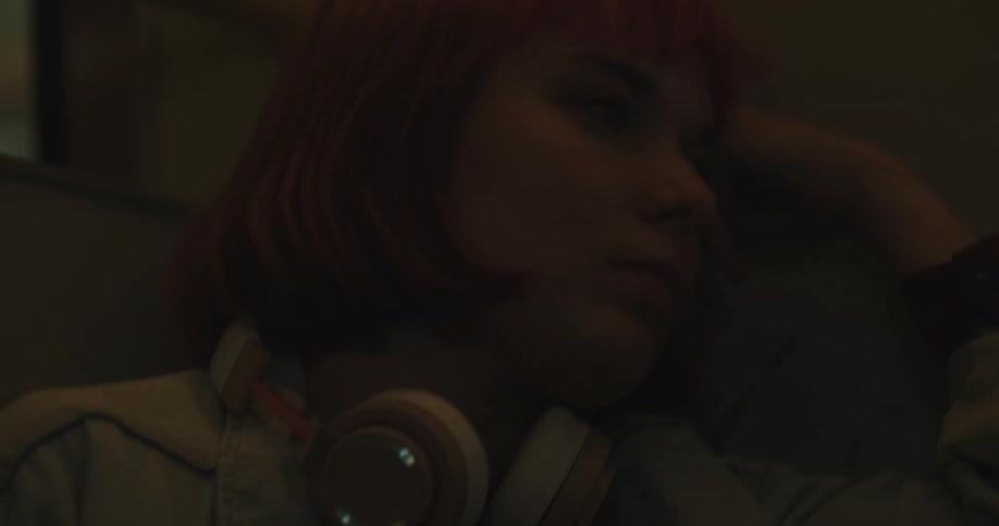 A teenage girl with pink hair and headphones around her neck is sleeping on a train